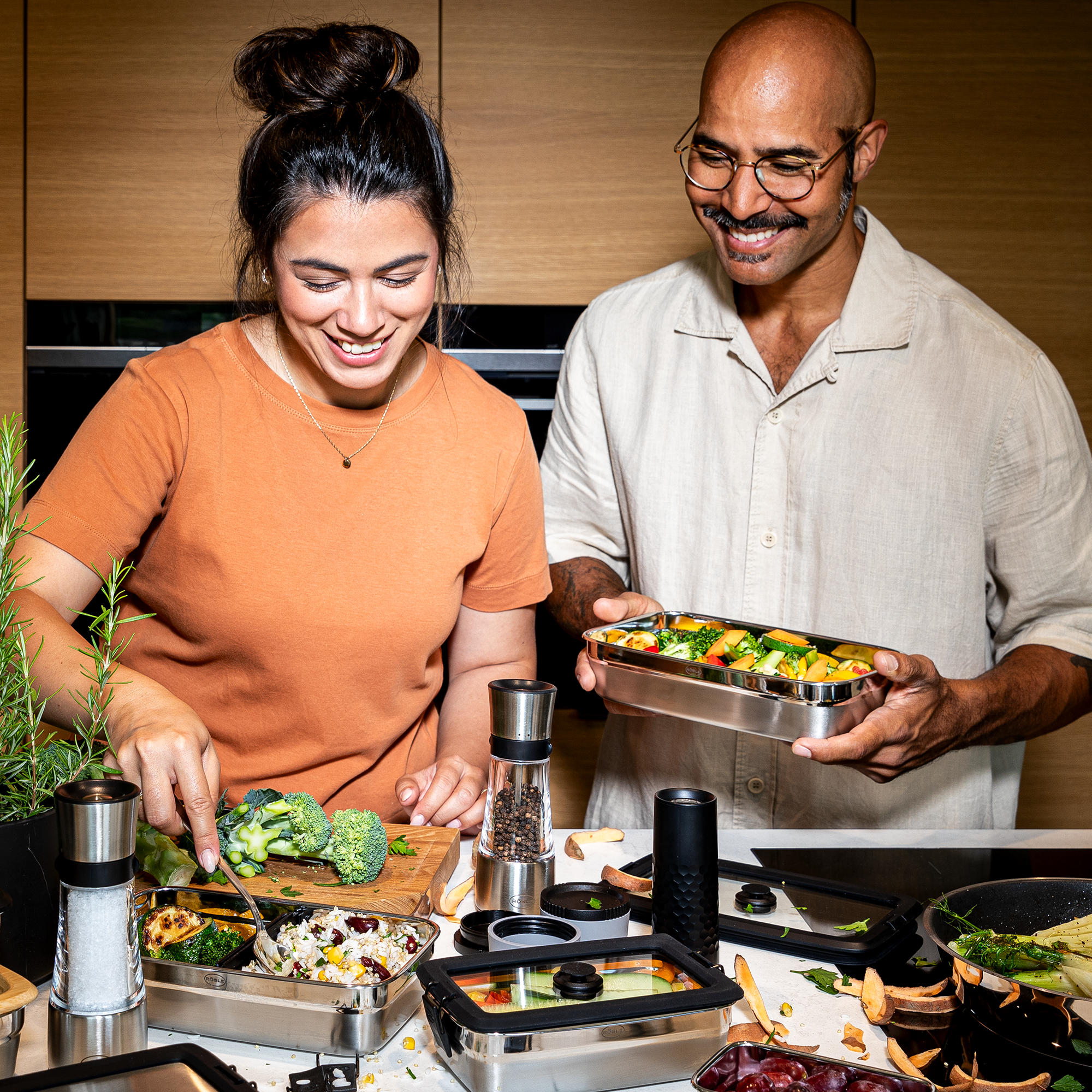 Zwei Personen beim Kochen in der Küche Zwei Personen in einer modernen Küche, eine Frau serviert Brokkoli, ein Mann hält eine Edelstahl-Behälter...