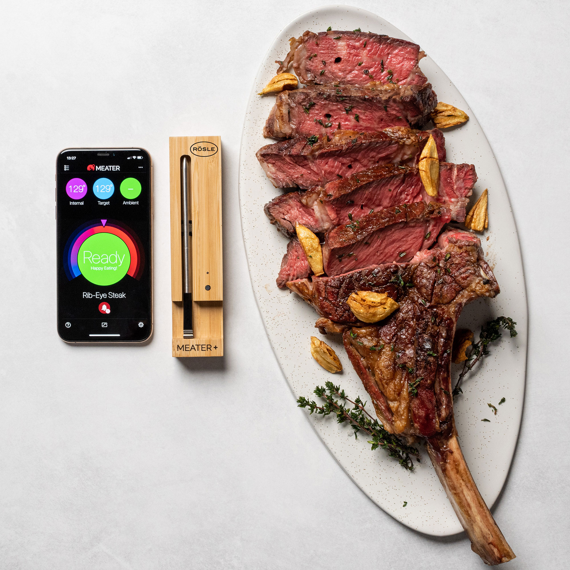 RÖSLE MEATER+ und gegrilltes Fleisch MEATER+ mit Smartphone-App, gegrilltes Fleisch auf einem Teller, Knoblauch und Kräuter.