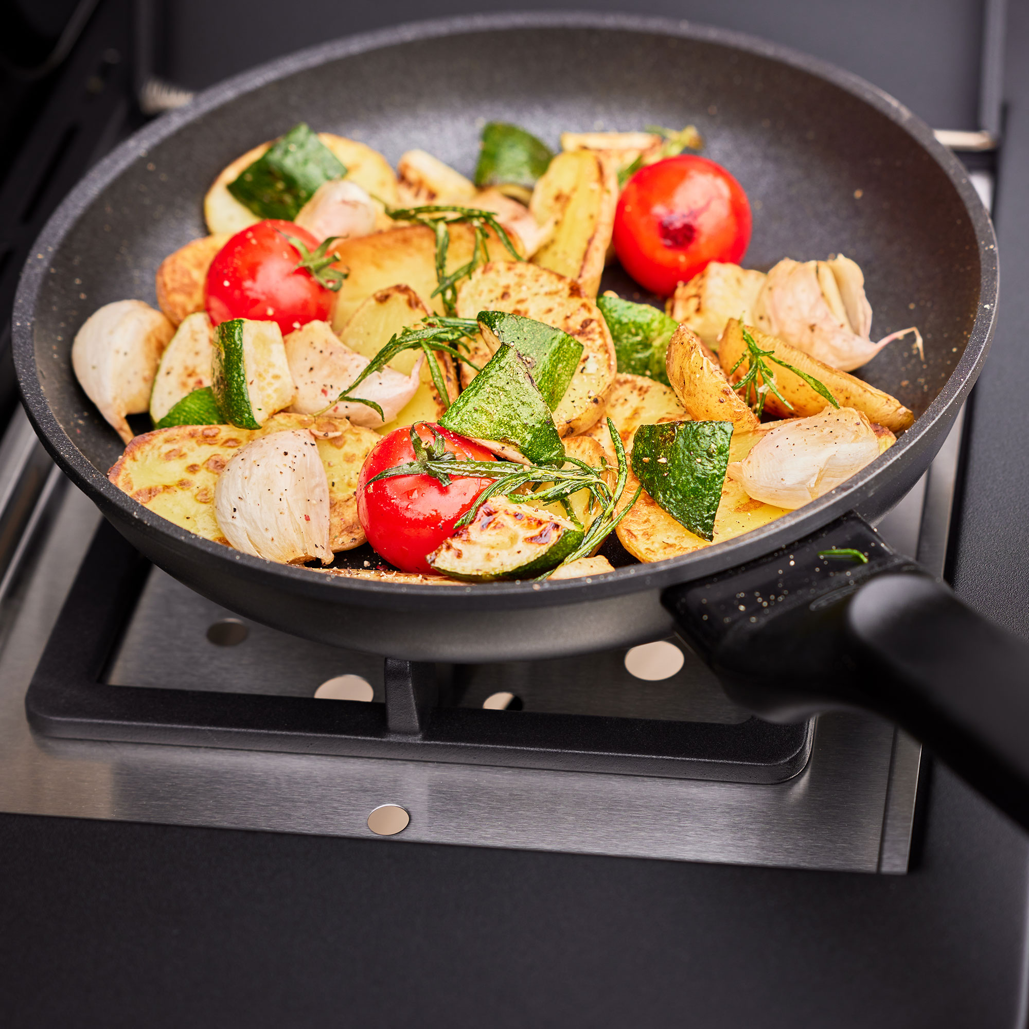 RÖSLE Gasgrill mit Pfanne und Gemüse Pfanne mit gebratenem Gemüse, darunter Zucchini, Tomaten und Kartoffeln, auf einem Gasgrill.