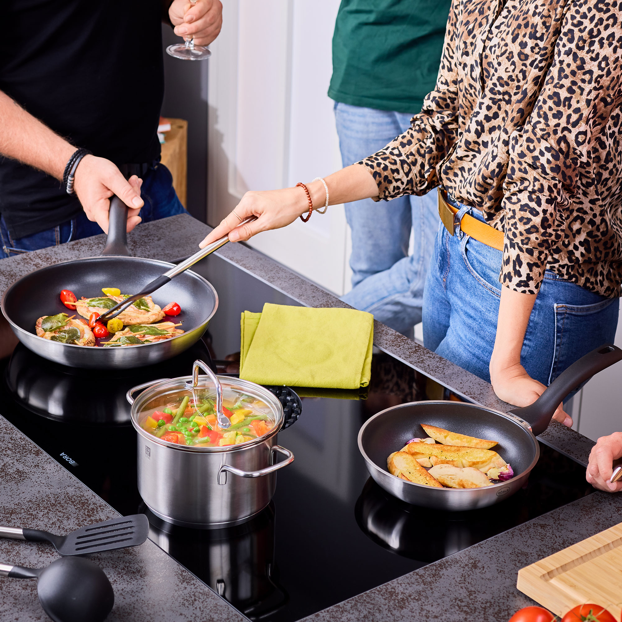 Pfannen-Set Basic Line in Aktion, mit Gemüse und Kartoffeln auf dem Herd, umgeben von Personen.