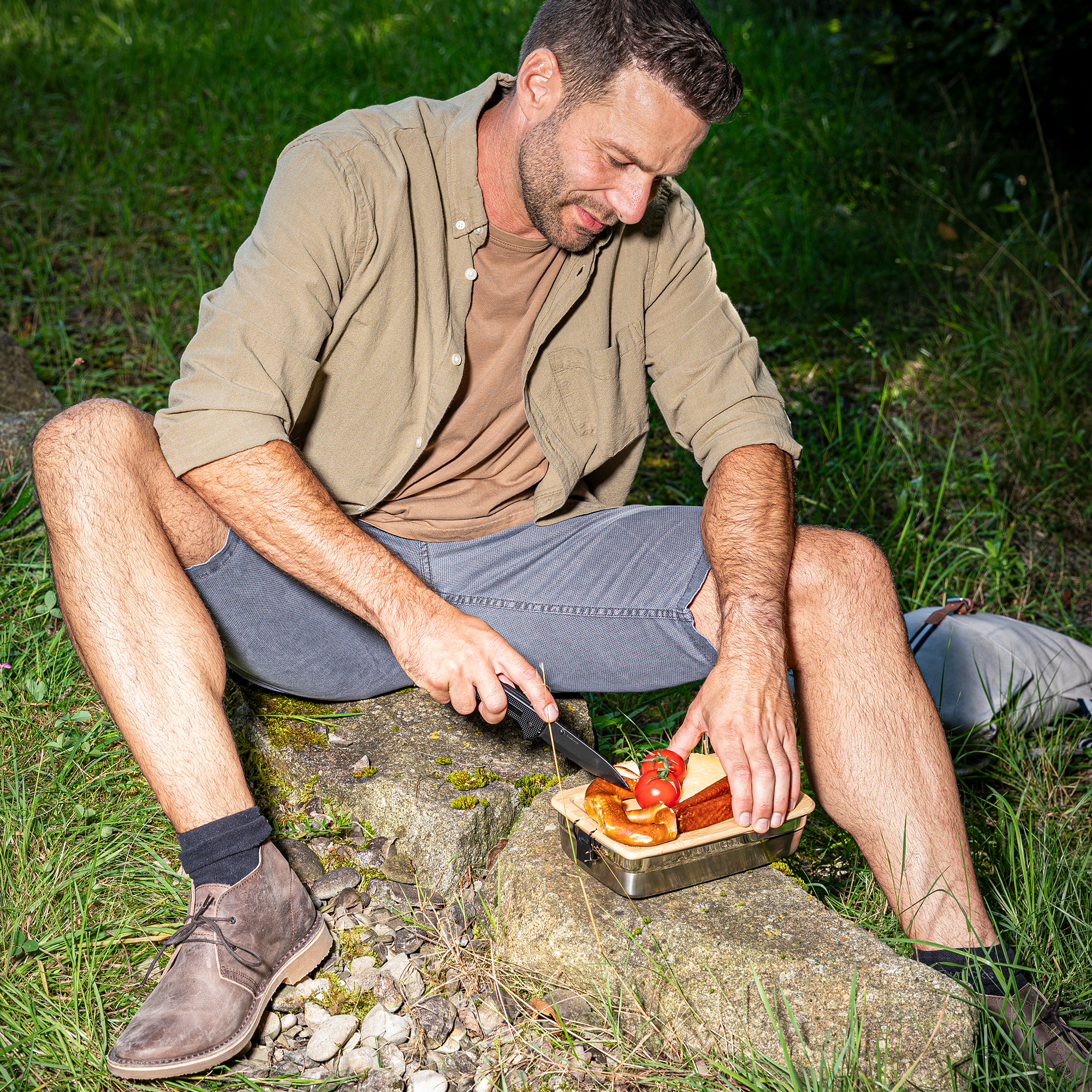 Mann schneidet Brot auf einem Stein im Freien, trägt ein khakifarbenes Hemd und Shorts.