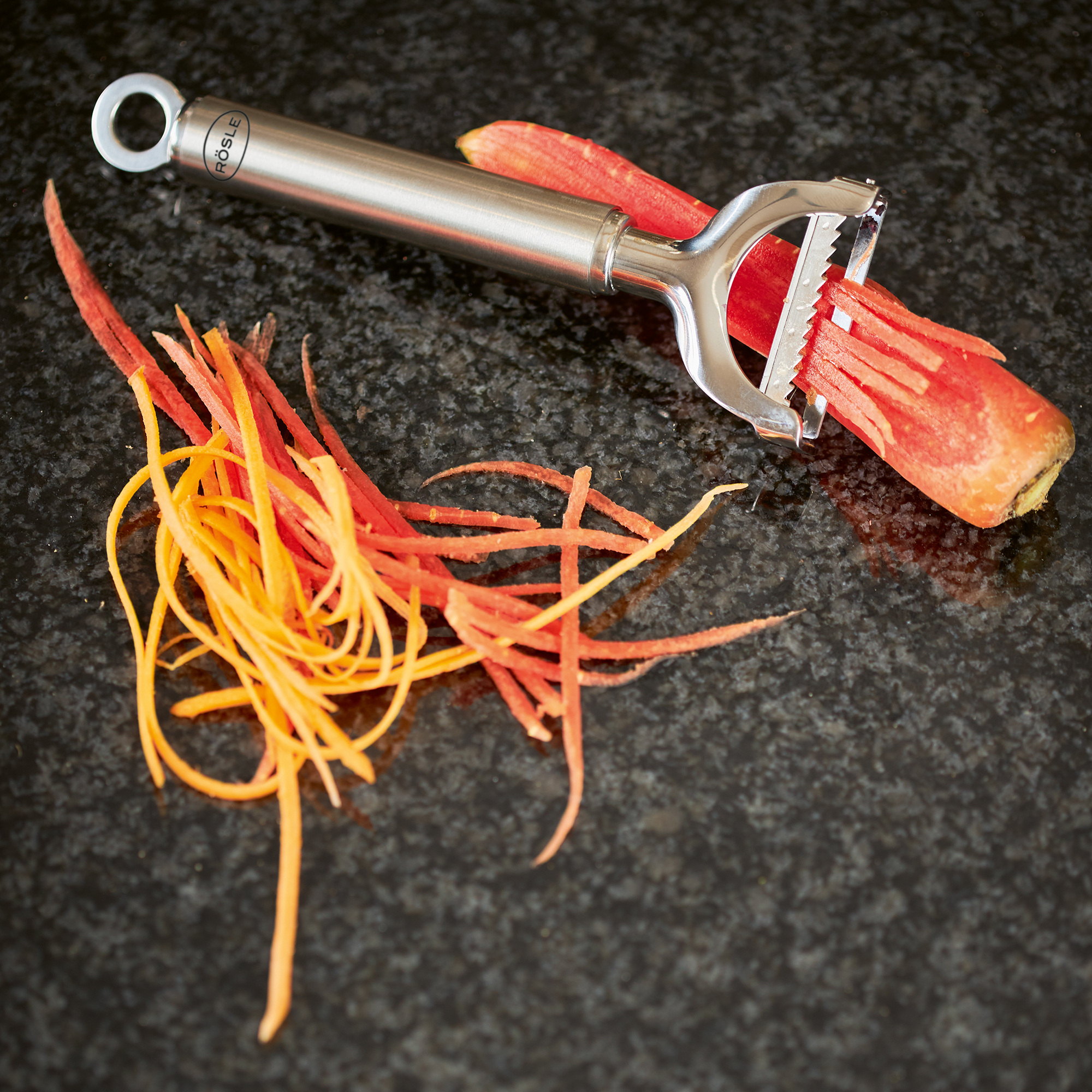 RÖSLE julienne peeler Stainless steel julienne peeler with carrot and apple strips on a black work surface.