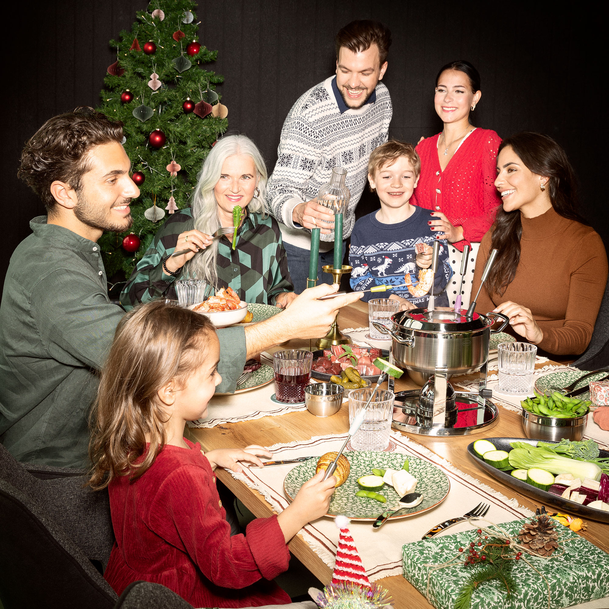 Familie und Freunde genießen ein Fondue-Essen an einem festlich gedeckten Tisch mit Weihnachtsbaum im Hintergrund.