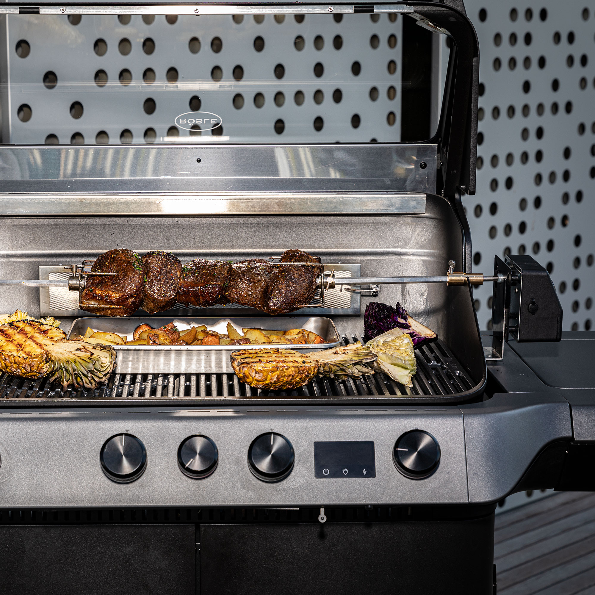 RÖSLE Drehspieß auf Grill Drehspieß mit Fleisch und Gemüse auf einem Grill, im Hintergrund eine moderne Grillumgebung.