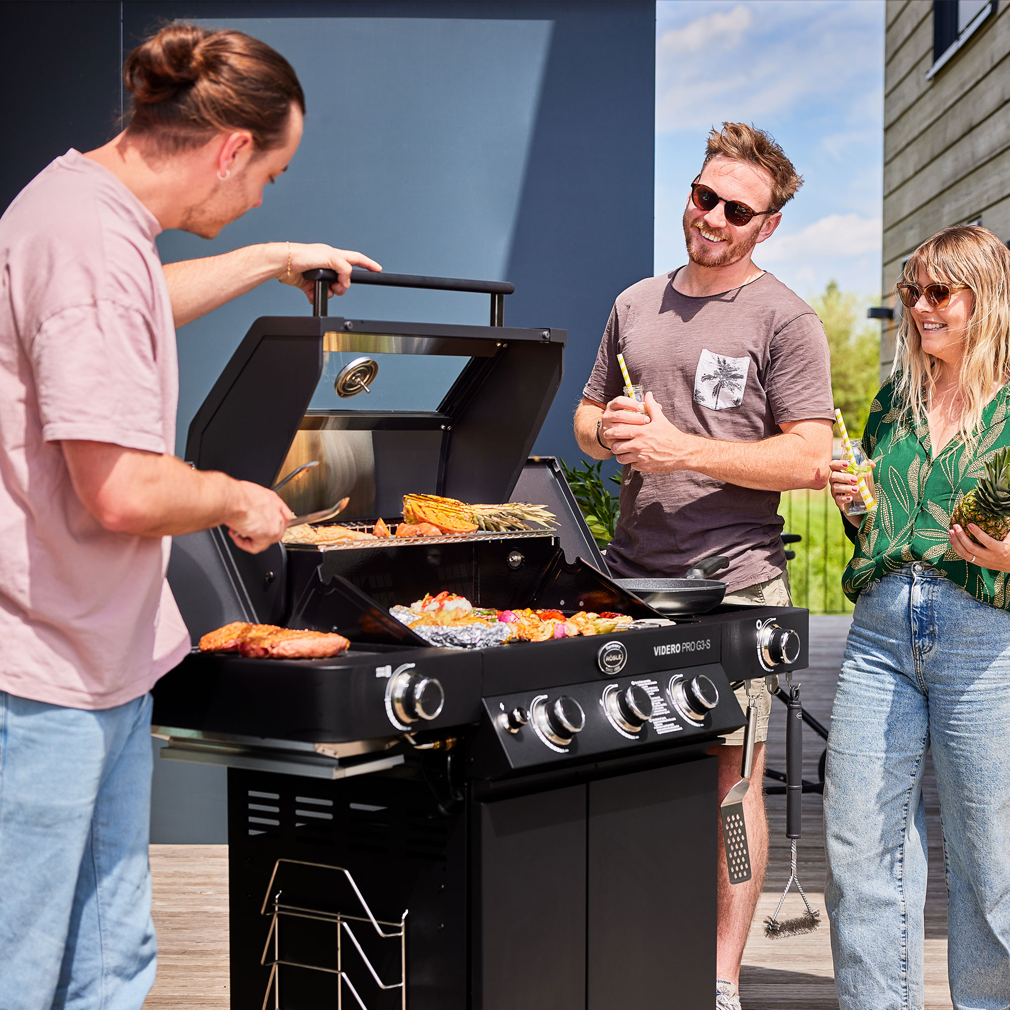 RÖSLE Gasgrill Videro G3-S PRO in Benutzung Gasgrill Videro G3-S PRO mit Grillgut, drei Personen stehen um den Grill, lächeln und unterhalten...