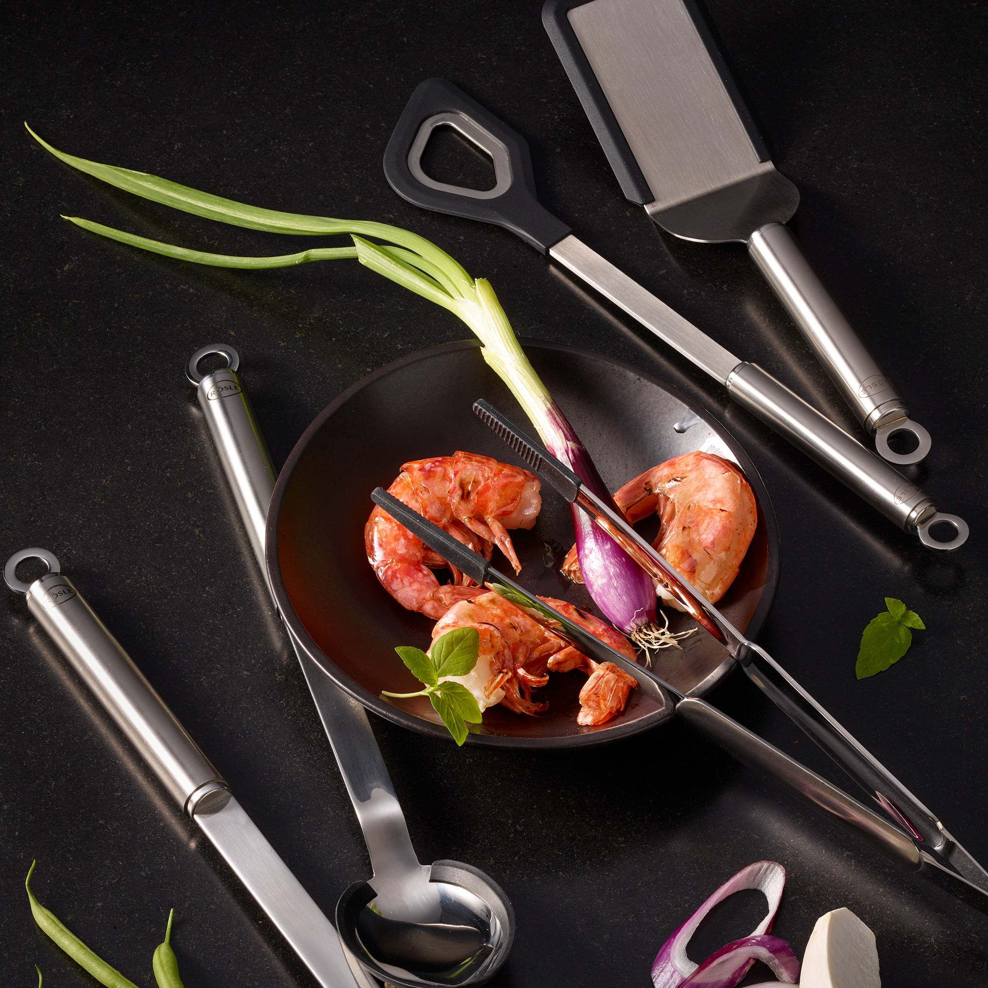 RÖSLE round handle cooking spoon and kitchen utensils Round-handled silicone cooking spoon with other kitchen utensils and ingredients on a black...