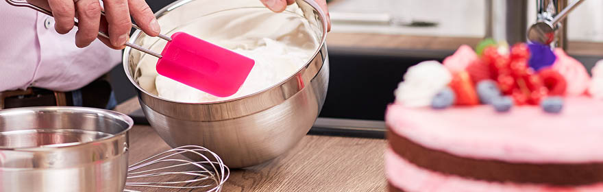RÖSLE mengkom met spatel en cake Roestvrijstalen mengkom met een roze spatel terwijl een persoon deeg roert. Een versierde...