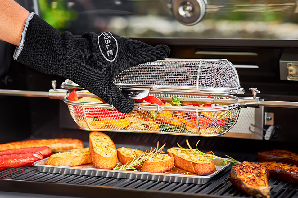 Rotating spit basket is opened with gloves. The rotisserie basket contains grilled vegetables