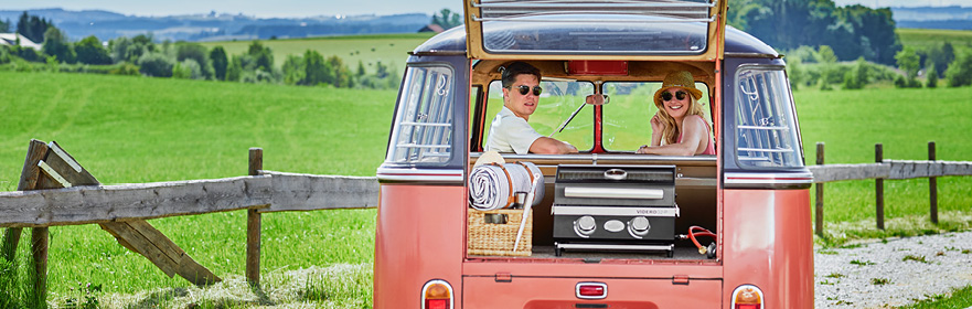 Paar schaut aus einem VW Bus mit  Gasgrill BBQ-Portable Videro G2-P im Kofferraum 