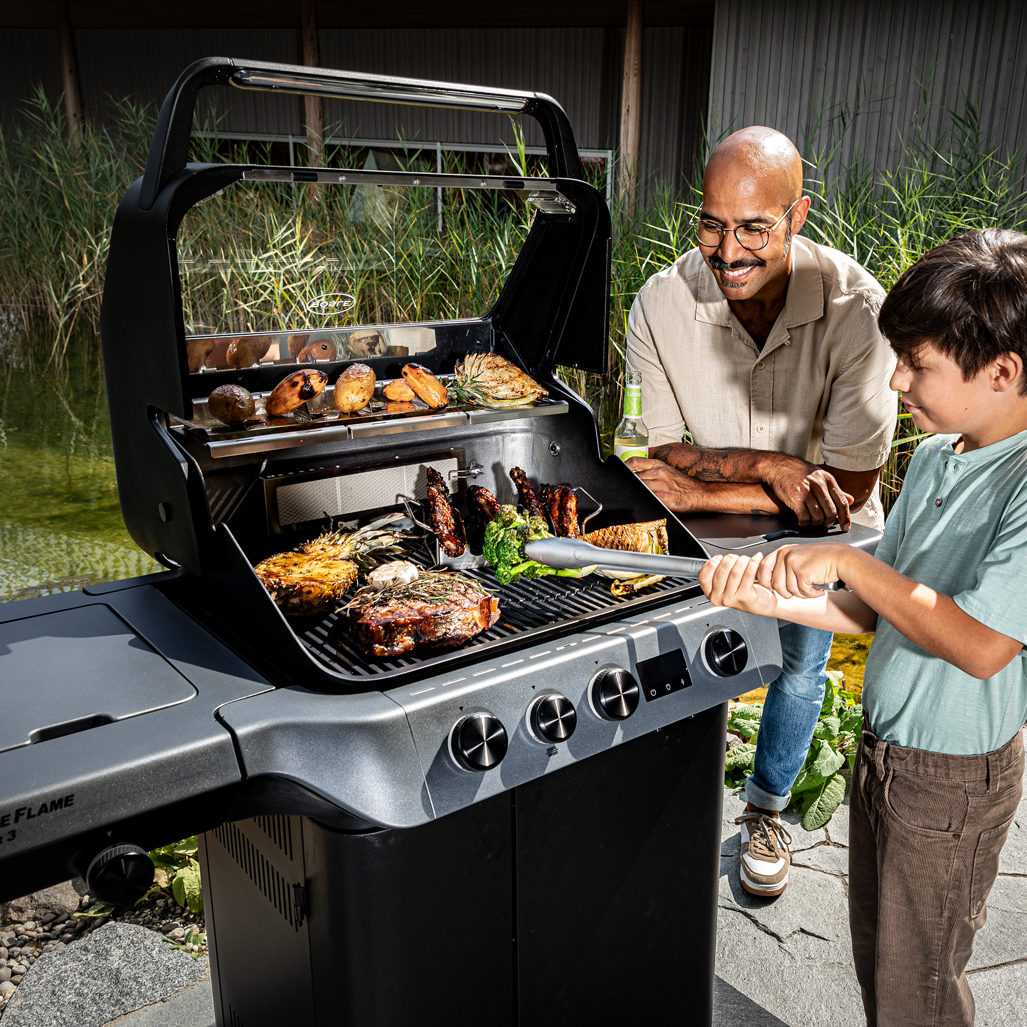 Blazeflame Master Grill mit gegrilltem Fleisch und Gemüse, ein Erwachsener und ein Kind beim Grillen.
