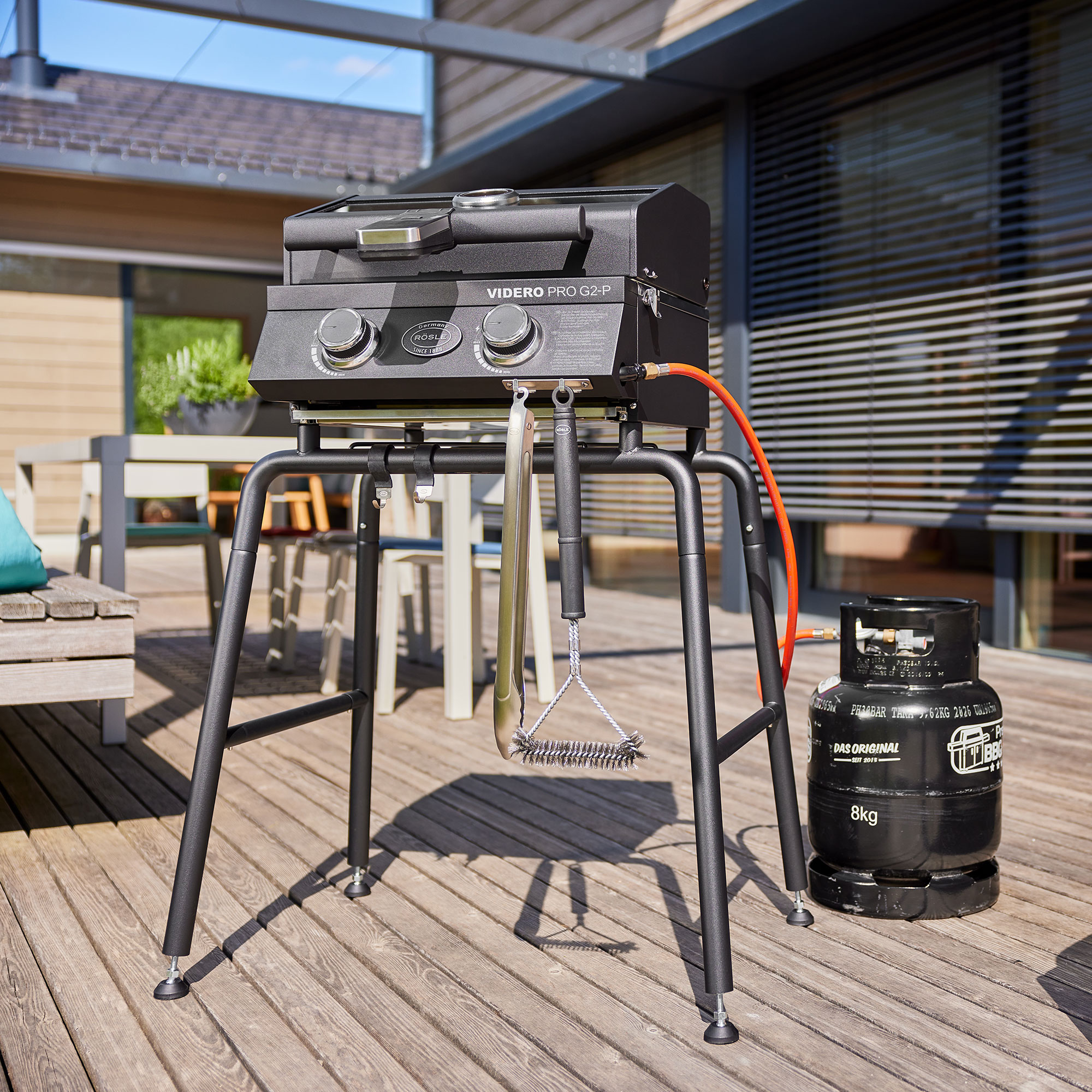 RÖSLE Videro G2-P Gasgrill auf Untergestell Videro G2-P Gasgrill auf einem stabilen Untergestell, mit Grillbesteck und Gasflasche daneben.