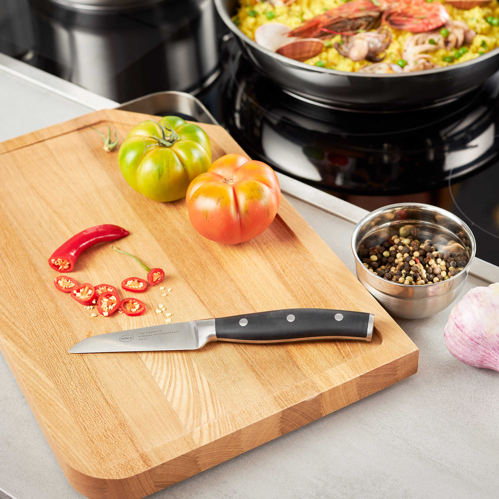 RÖSLE Schälmesser auf Holzbrett mit Gemüse Schälmesser, 8 cm, mit schwarzem Griff, auf einem Holzbrett neben frischem Gemüse und Gewürzen.