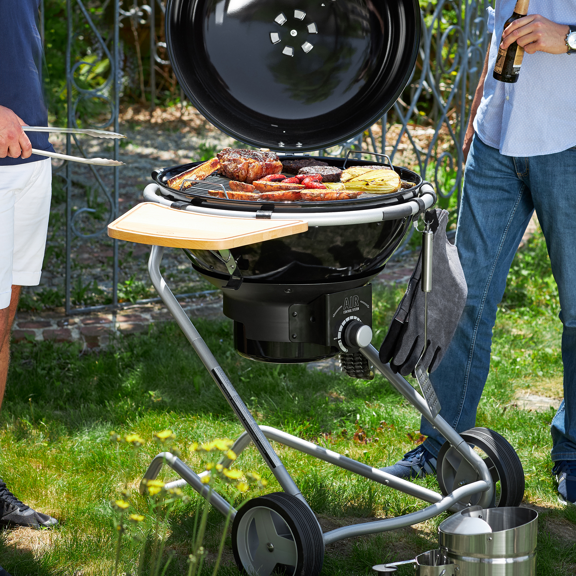 RÖSLE Grill im Freien mit Grillgut Grill mit geöffnetem Deckel, Fleisch und Gemüse auf dem Rost, zwei Personen stehen daneben,...