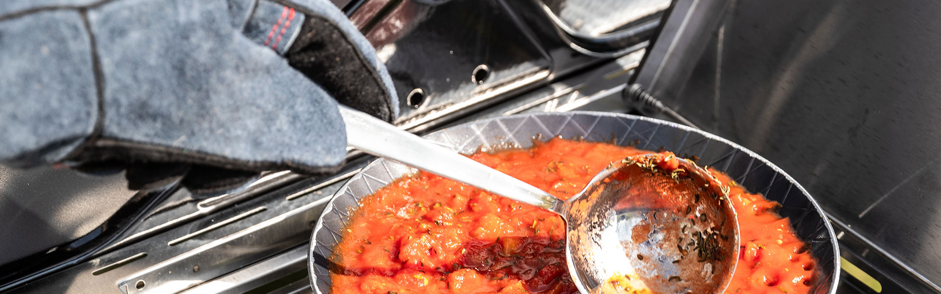 Grillzubehör mit Tomatensauce Grillzubehör: Jemand schöpft Tomatensauce mit einer Kelle aus einer Grillpfanne.