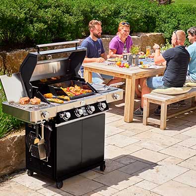 Barbecue with open lid in the foreground and behind it 4 friends sitting together at the table and eating.