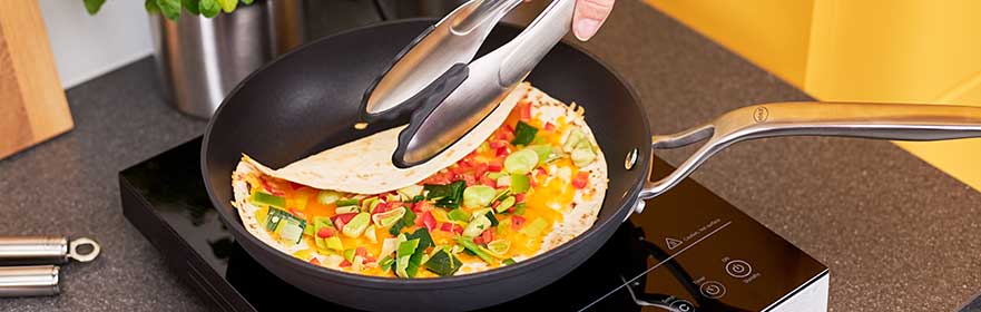 RÖSLE pan with vegetables and tortilla Pan with colourful vegetables and tortilla, tongs in hand for turning, on a hob.