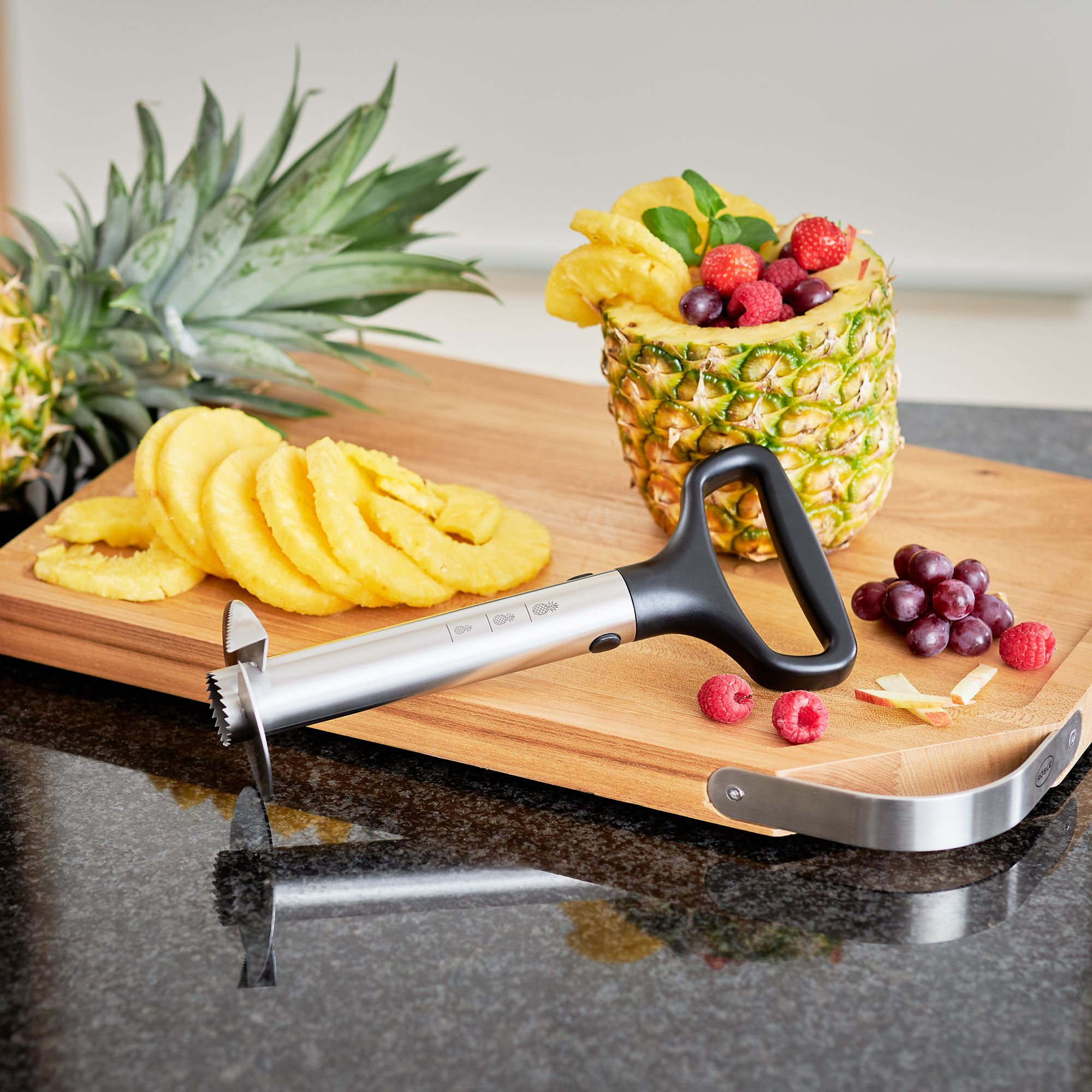 RÖSLE pineapple slicer on wooden board with fruit Stainless steel pineapple slicer with black handle, next to sliced pineapple and a pineapple...